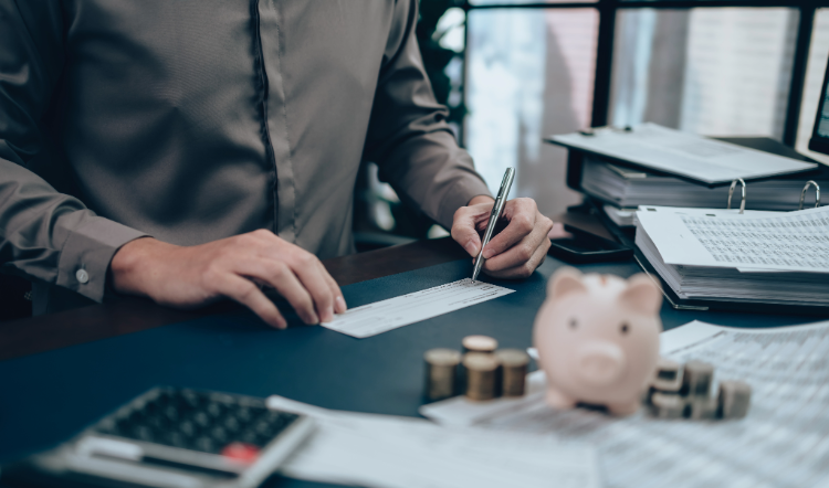 Man sitting at a desk is writing on a check with a piggy bank and change in the foreground.
