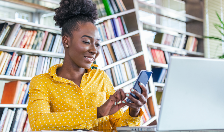 A woman in a bright yellow sweater looks at her smartphone and also has a laptop open in front of her.